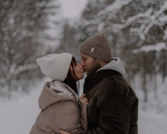 A couple shares a romantic kiss on a snowy forest path during winter, surrounded by conifer trees.